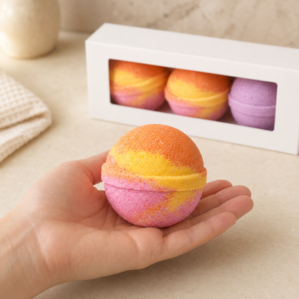 A hand holds a round bath bomb with a pink, yellow, and orange marbled pattern in the foreground. In the background, a white box containing three more bath bombs is visible on a light beige countertop.