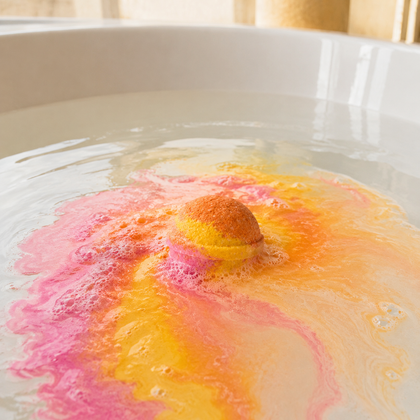 A spherical bath bomb dissolves in clear water inside a white bathtub, creating a colorful splash of pink, orange, and yellow.