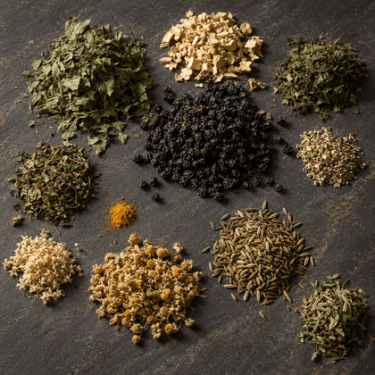 An overhead flat-lay arrangement of various loose-leaf herbal teas and spices on a dark textured slate background, including green tea, chamomile, black seed, cumin, turmeric, and dried roots.
