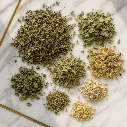 Seven different loose piles of dried and fresh herbs, flowers, and botanicals arranged on a white marble surface with a gold edge.
